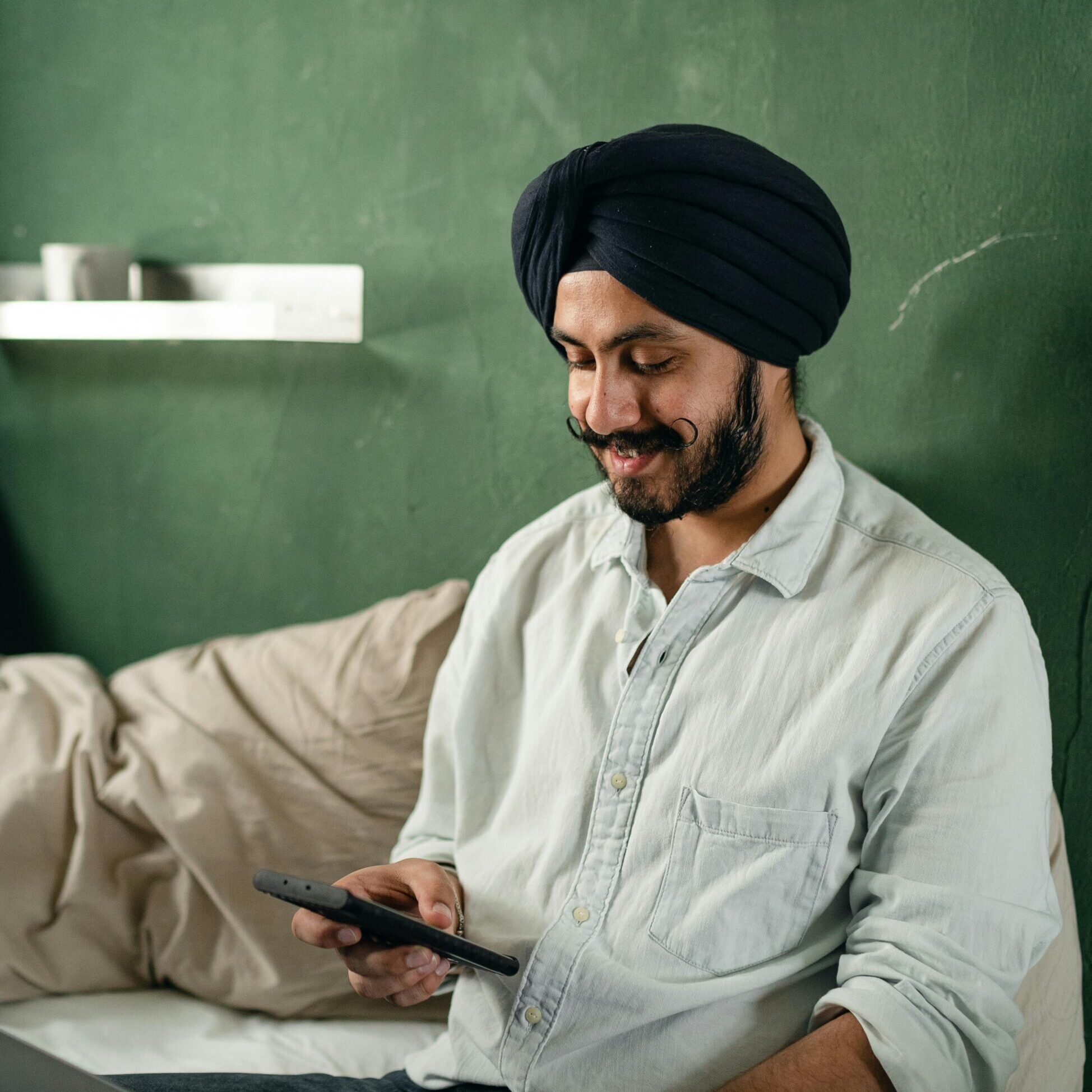 Cheerful bearded Indian male wearing turban sitting with legs crossed on comfortable bed with laptop on laps and browsing smartphone